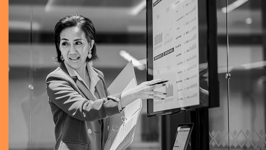 A person in a business suit points at a large screen displaying various charts and graphs during a presentation.