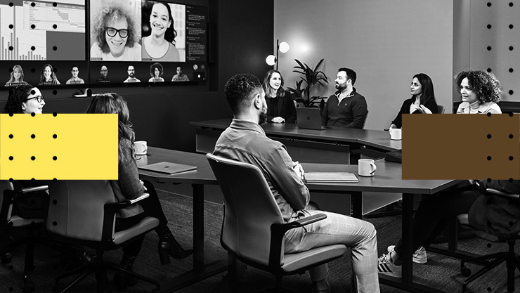 A conference room with people seated at a table, participating in a video conference displayed on large screens.