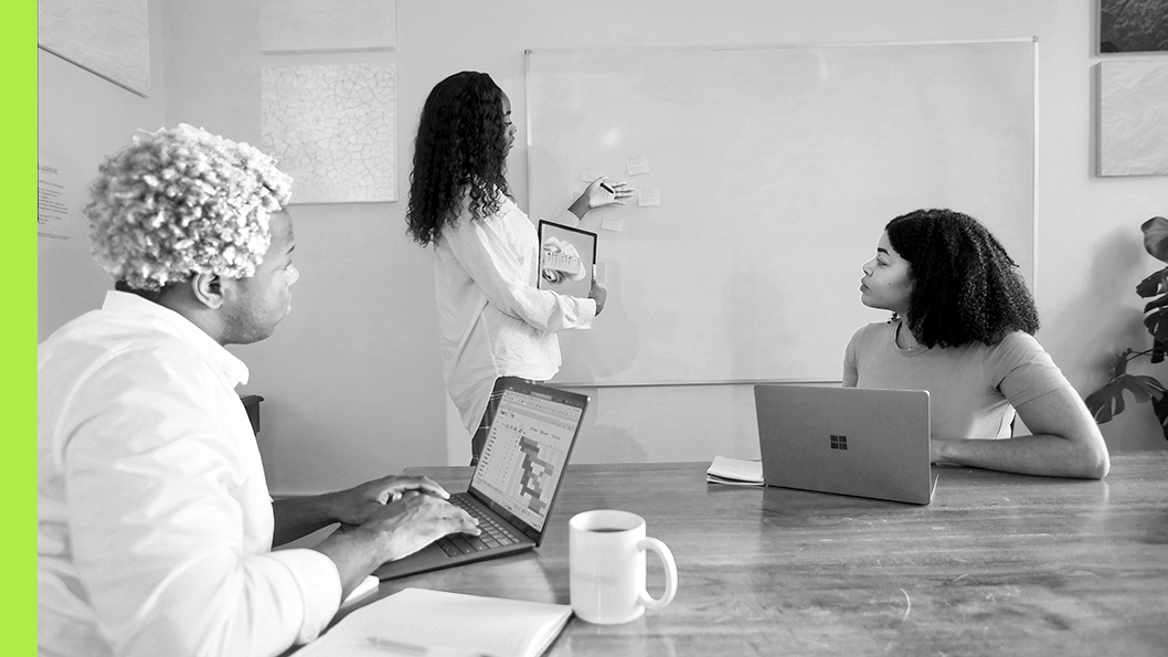 One person presents sticky notes on a whiteboard while another, seated at a laptop, follows along.