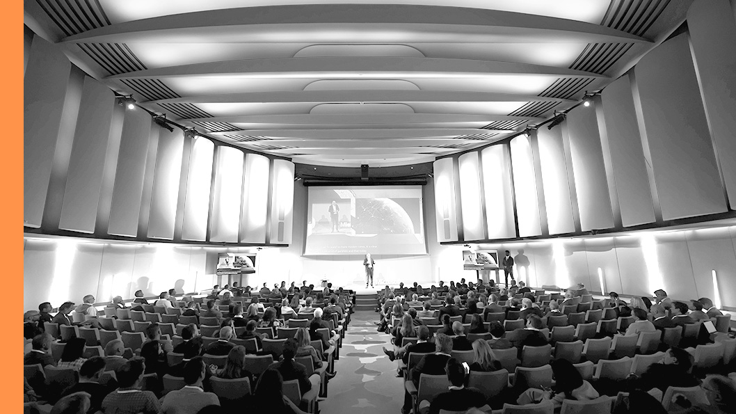A wide view of a large auditorium filled with seated attendees facing a stage and presentation screen.